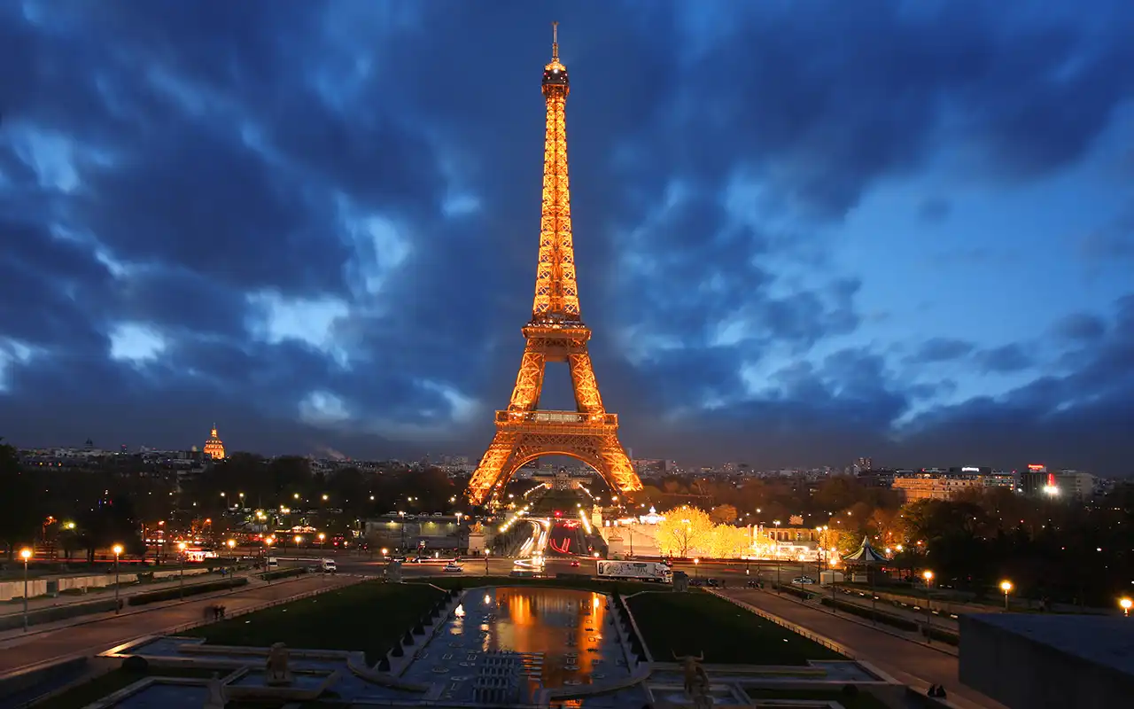 Majestic Eiffel Tower in Paris, France lit up at dusk with a cloudy blue sky backdrop.